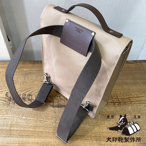 犬印純綿帆布製直立式側背包，淺褐色帆布包身，配有深棕色皮革提把、皮革裝飾片及肩帶，肩帶為深灰色編織材質，可轉換為後背包使用。