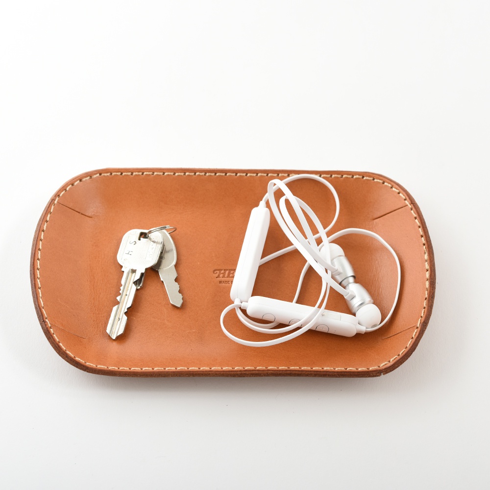 職人手作的淺棕色皮革收納盤，盤中放著一串鑰匙和一組白色無線耳機，皮革邊緣縫線清晰。