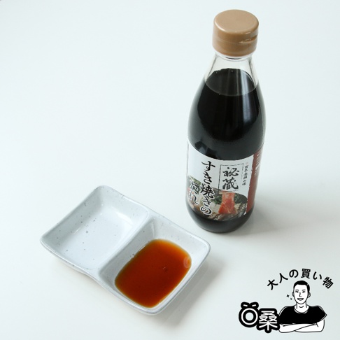 一瓶深褐色醬汁的玻璃瓶，瓶蓋為淺棕色，瓶身標籤上有日文文字及壽喜燒相關圖案。旁邊有一個白色雙格醬料碟，其中一格盛有少量深紅色醬汁。