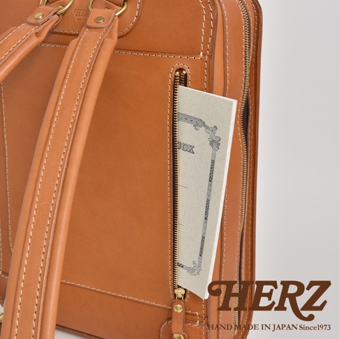 職人手作皮革商務電腦後背包 R-25，展現經典棕色皮革材質，車縫線清晰。背包側面有一個拉鍊口袋，露出部分筆記本。