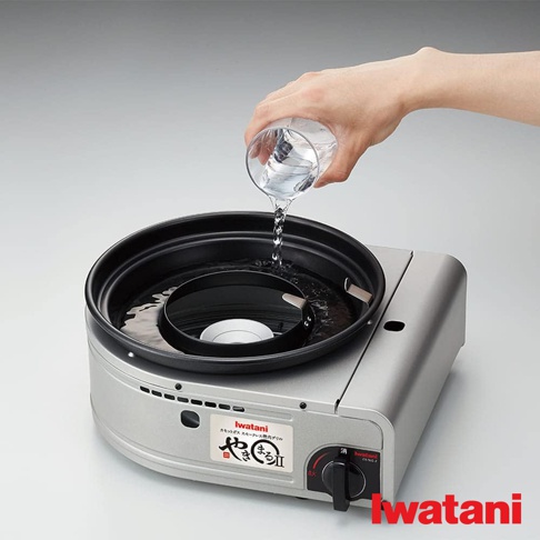銀色金屬外殼的 Iwatani 無煙燒烤爐，上方黑色烤盤正在注入清水，烤爐側面有品牌標誌和控制旋鈕。