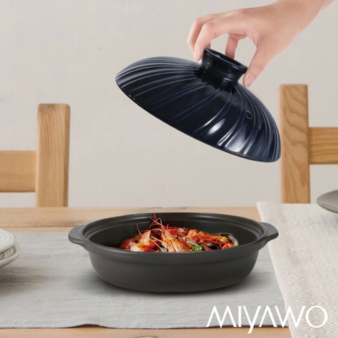 黑色直火系列陶板鍋，鍋身為啞光質地，盛有鮮蝦及配料。一只手正提起一個帶有螺旋紋路的黑色陶製鍋蓋。