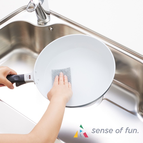 一隻手拿著白色平底鍋，另一隻手用灰色鬆餅紋海綿巾擦拭鍋內。背景為不鏽鋼水槽和水龍頭。