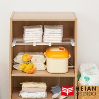 日系木紋風縫隙置物架，層架上擺放兩疊尿布、尿布墊、可愛的黃色小鴨玩偶、橙色帶蓋的儲物罐，下方層架擺放疊好的嬰幼兒衣物、襪子，以及印有平安伸銅 Logo 的籃子，整體收納整齊。