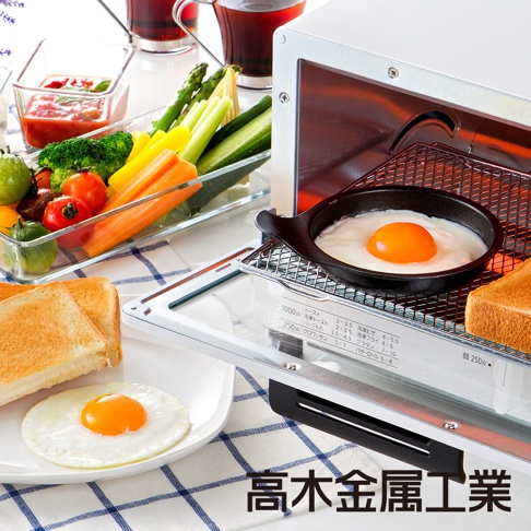 日本製烤箱內，一個黑色圓形烤盤盛著一個金黃蛋黃的煎蛋，烤箱架上擺放著烤過的吐司。盤邊有蔬菜拼盤，包含小番茄、綠花椰、黃甜椒、紅蘿蔔、蘆筍和黃瓜。