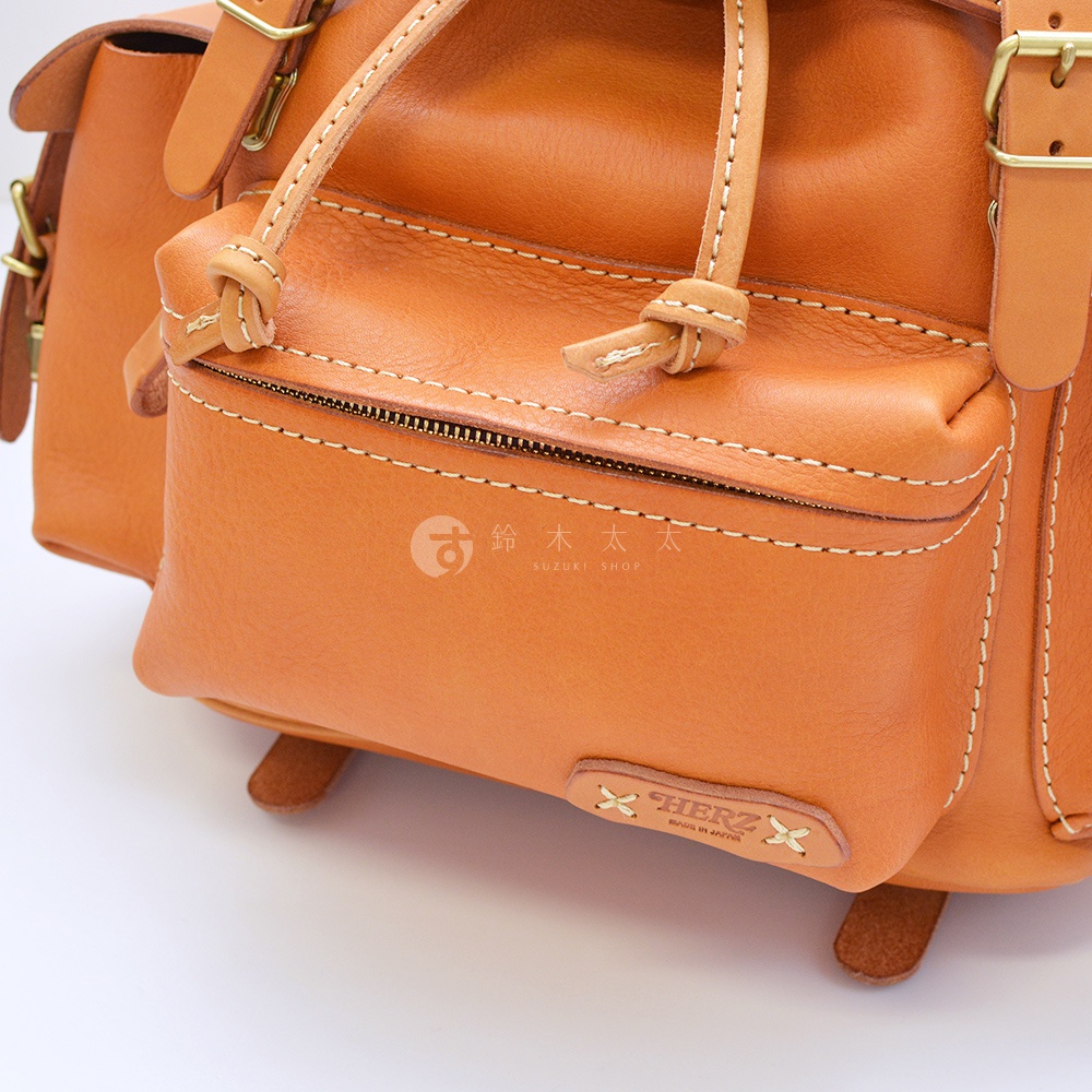 職人手作皮革復古風旅行後背包 R-65，特寫展示棕褐色皮革材質、精緻車縫線、拉鍊及束繩細節，右下角有品牌標誌。