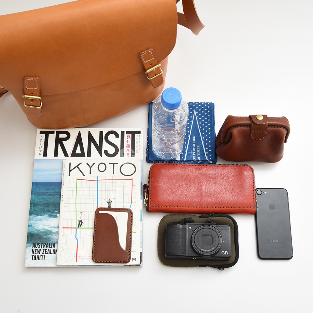 多種皮革小物與雜誌，包含一個棕色郵差包、一個棕色零錢包、一個紅色長夾、一個手機，以及一本標示有TRANSIT KYOTO字樣的雜誌。