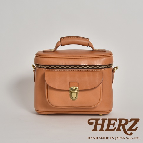 職人手作的淺棕色皮革相機包，圓潤的包身設計，頂部有提把，正面有一個翻蓋式小口袋，附有金色金屬鎖扣，整體呈現簡約復古風格。