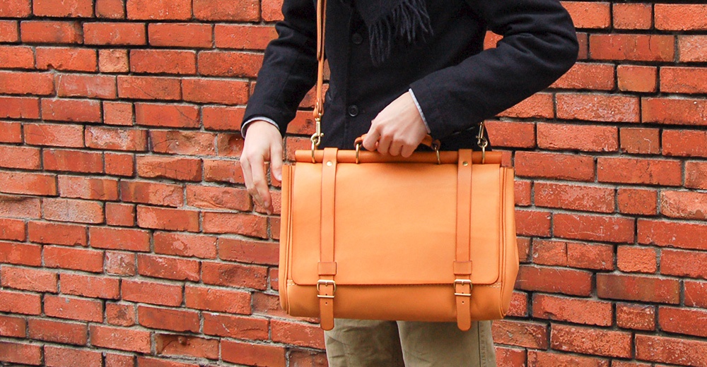 職人手作的橘色皮革側背包，搭配黑色外套，拎在磚牆前。