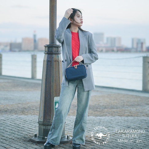 穿著灰色西裝外套、紅色內衣和牛仔褲的女性，手提一個藍色斜背包，站在路燈旁。