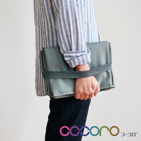 手持一個綠灰色、折疊起來的購物車，車身由布料製成，並以彈性織帶固定。