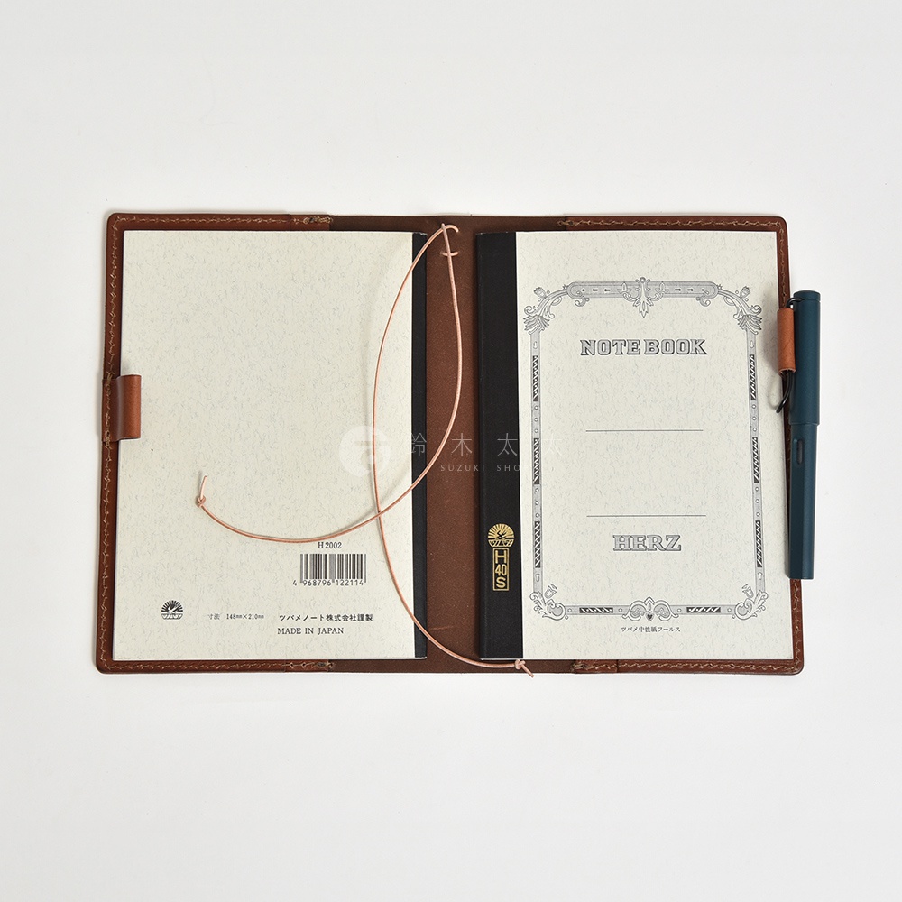打開的棕色皮革手帳書套，內有兩本筆記本，一本為空白，另一本則有裝飾性邊框和筆記本字樣，右側夾著一支綠色鋼筆。