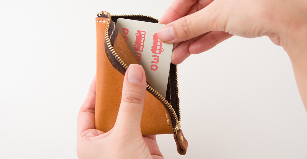 雙手拿著一個日本HERZ職人手作的橘棕色皮革迷你錢包，錢包內部襯有深色皮革，右側正從錢包開口處取出印有紅色交通工具圖案的票卡，展現精緻車縫線細節與皮革質感。