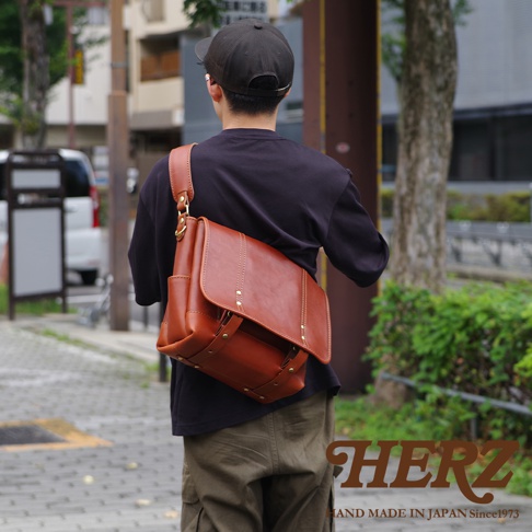 棕色皮革復古郵差包，附有肩帶和金屬扣。包身兩側有側袋，正面有翻蓋和裝飾性鉚釘。