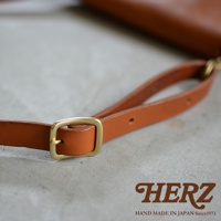 HERZ 品牌標誌，下方為 HAND MADE IN JAPAN Since1973 字樣，圖片聚焦於棕色皮革斜背包的肩帶細節，可見黃銅色金屬調節環，整體風格呈現手工質感。