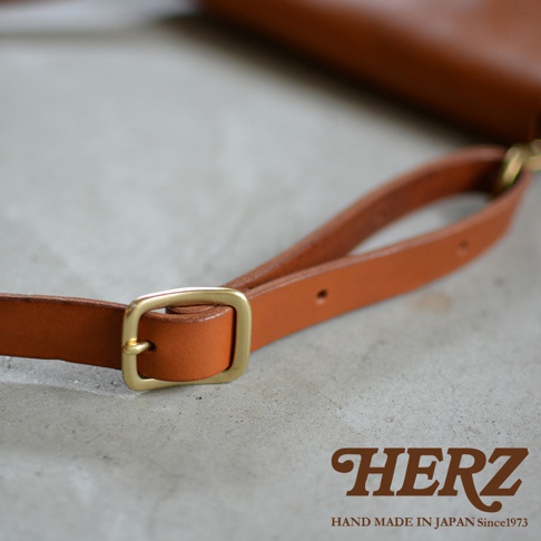 HERZ 品牌標誌，下方為 HAND MADE IN JAPAN Since1973 字樣，圖片聚焦於棕色皮革斜背包的肩帶細節，可見黃銅色金屬調節環，整體風格呈現手工質感。