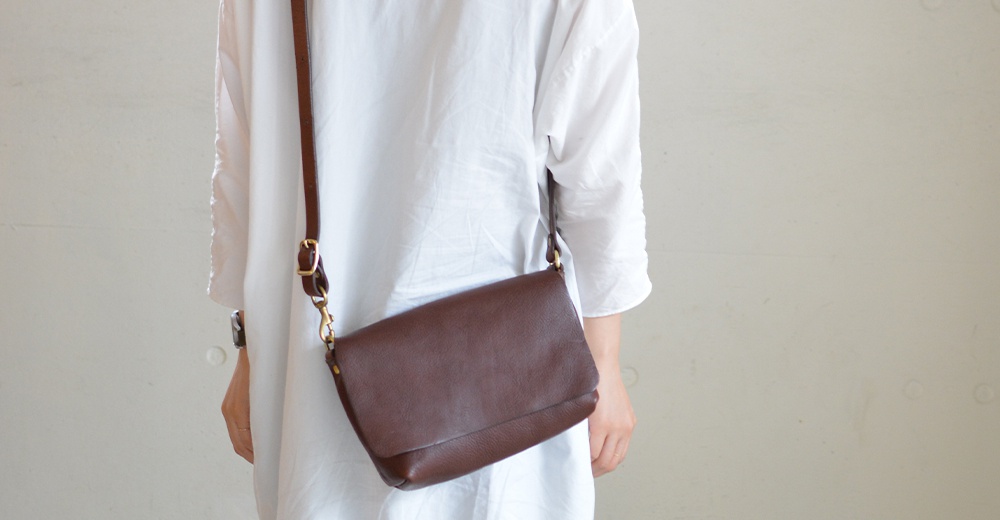 一位身穿白色襯衫的人斜背著一個深棕色皮質翻蓋包，包包有著自然的紋理和金屬扣環。
