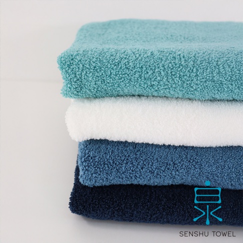 一疊四條envelopper迷你浴巾，顏色由上至下為薄荷綠、純白、海軍藍與深藍。浴巾材質蓬鬆，觸感柔軟。