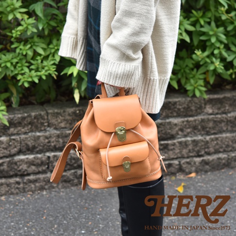 一位穿著米色針織外套的女性，手持日本HERZ職人手作的經典迷你皮革後背包，背包呈現溫潤的淺棕色調，具備金屬扣環與束口設計，整體展現簡約質感的手工皮革工藝風格。