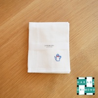 白色八重紗精繡擦拭巾，織有網格紋理，左下方繡有藍色與紅色線條點綴的茶壺圖案，產品標籤為白色，印有日文及英文產品名稱。