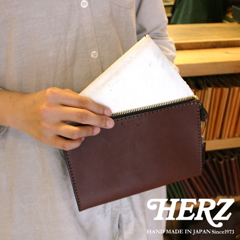 職人手作皮革三角底拉鍊手拿包，深棕色皮革，金色拉鍊，包面上壓印有「HERZ」品牌標誌。
