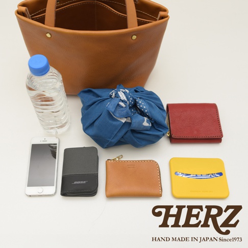 職人手作的駝色皮革托特包，內有手機、灰色卡夾、棕色零錢包、紅色皮夾，以及藍色印花布包裹的物品，還有黃色小物件。