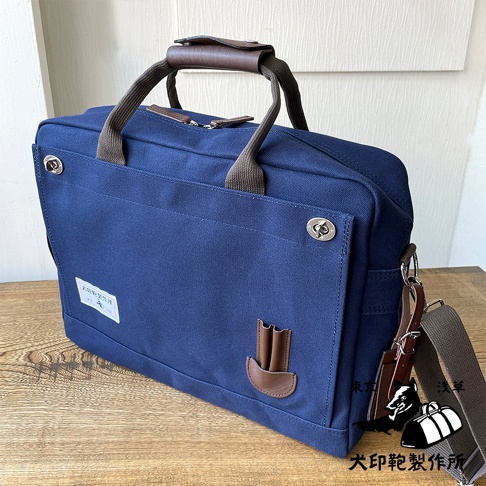 深藍色純綿帆布商務公事包，B4尺寸，配有棕色皮革提把和可拆卸肩帶，正面有金屬扣環和品牌標籤。