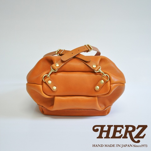 職人手作皮革輕旅行後背包，採用質感棕色皮革，設有兩條可調節的皮革提把，搭配金色金屬扣環和鉚釘，整體造型簡約實用。