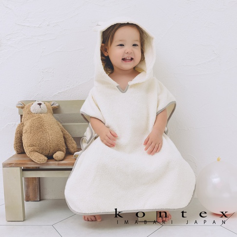 一位穿著白色斗篷式連帽浴巾的嬰兒，浴巾帶有可愛的泰迪熊耳朵設計，邊緣為灰色滾邊。