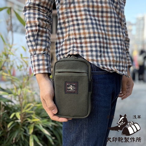 一位穿著格紋襯衫與牛仔褲的男子，側身展示繫於腰間的軍綠色犬印鞄製作所帆布手機包，包身正面縫有品牌標籤，右下角亦有標誌性的犬型圖案與文字設計。