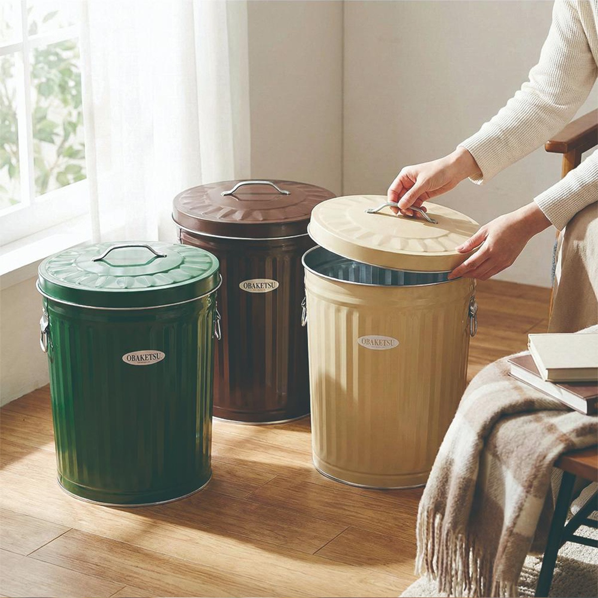 OBAKETSU 品牌三個不同顏色的日本製手工垃圾桶，其中一個米色款的蓋子被打開，旁邊還有一個裝有格紋毛毯的椅子。