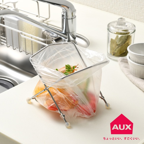 銀色金屬製的折疊式垃圾架，架著一個透明塑膠袋，袋內裝有廚餘，例如紅蘿蔔、番茄和香草。垃圾架有四個支腳，末端有白色塑膠墊。背景是廚房水槽和櫥櫃。