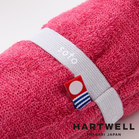 粉紅色毛巾捲起，繫有白色束帶，束帶上有 soto 字樣，一旁另有紅色小標籤，下方印有 HARTWELL MABARI JAPAN 字樣