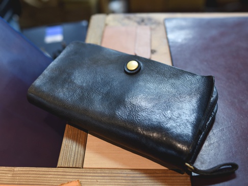 黑色皮革長夾，設有金色按扣和可拆卸腕帶，質地柔軟，適合旅行收納護照與機票。