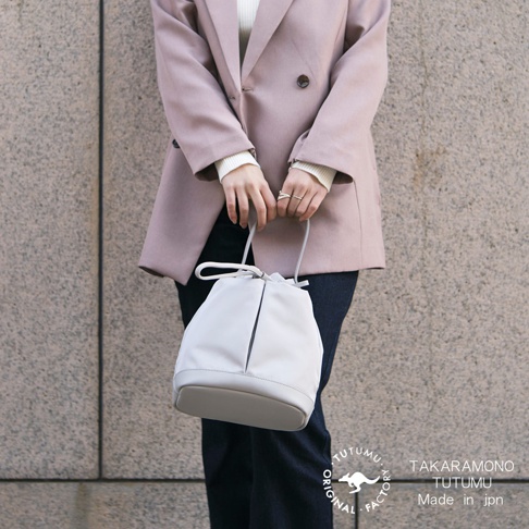 粉色西裝外套搭配白色內搭，手持一個灰白色抽繩水桶包，包包上有「TAKARA MONO TUTUMU」和「MADE IN JPN」字樣