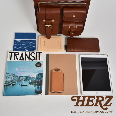 職人手作皮革三口袋三用後背包，搭配藍色印花手帕、雜誌、筆記本、皮革鑰匙包、長夾及平板電腦，整體為棕褐色調。