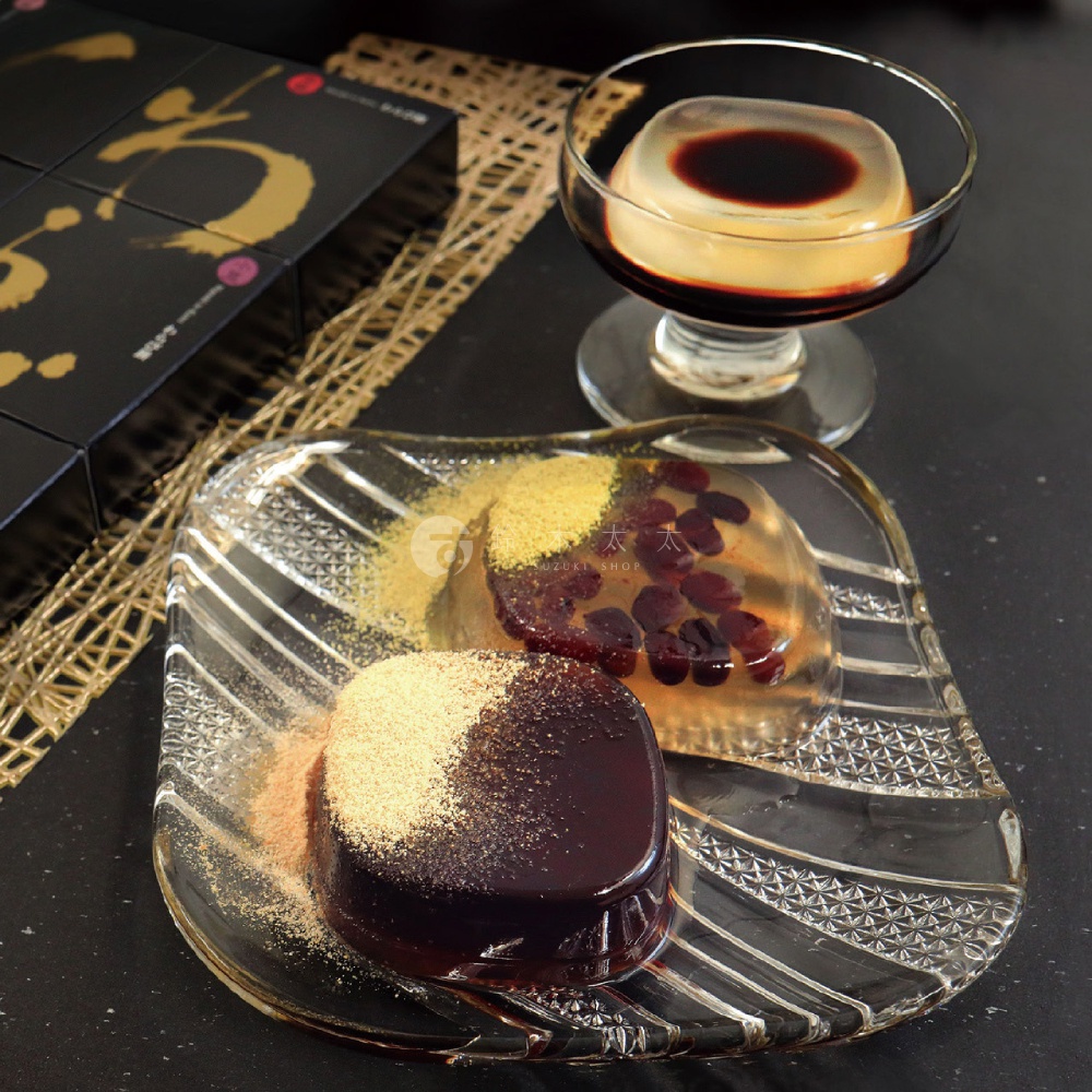 擺放在黑色桌面上的一組和風甜點，包括一個裝有深色糖漿的透明玻璃杯，以及一個刻花透明玻璃盤，盤上放置了一塊撒上黃豆粉的深色葛餅，和一塊內含紅豆的透明蕨餅，背景左側可見黑色包裝禮盒。