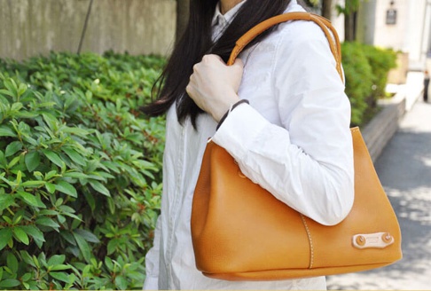 一位女士身穿白色襯衫，肩背一個焦糖色皮革托特包，包包有著簡潔的線條和明顯的縫線細節，以及一個淺色皮革的裝飾小牌。