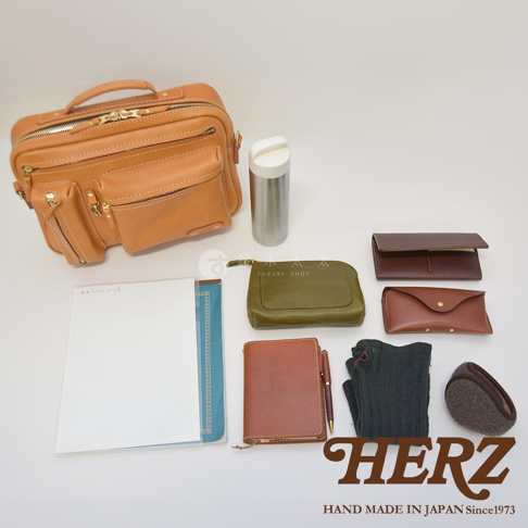 職人手作皮革兩用單肩包，呈現天然皮革的淺棕色，包身設計多個拉鍊口袋和翻蓋口袋。周邊配件包含軍綠色皮革小包、棕色長夾、棕色眼鏡盒、筆記本、鋼筆、灰色襪子與棕色毛絨耳罩。