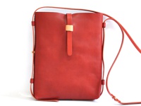 紅色皮革製成的直式單肩包，包身簡潔，正面中央有一條皮質束帶，束帶上附有淺棕色皮革裝飾，肩帶為細長皮質，兩側有金屬扣件。