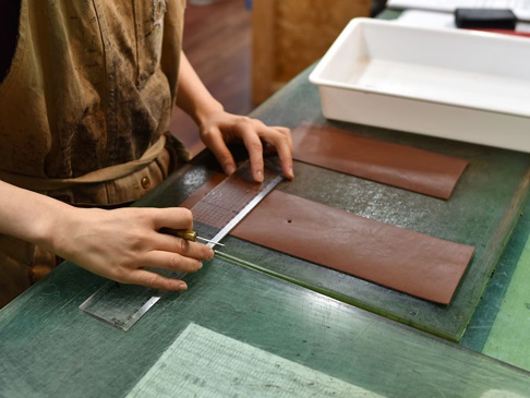 職人正在使用裁刀和尺，在深棕色皮革上進行切割。皮革表面有細微紋理，與綠色工作檯形成對比。