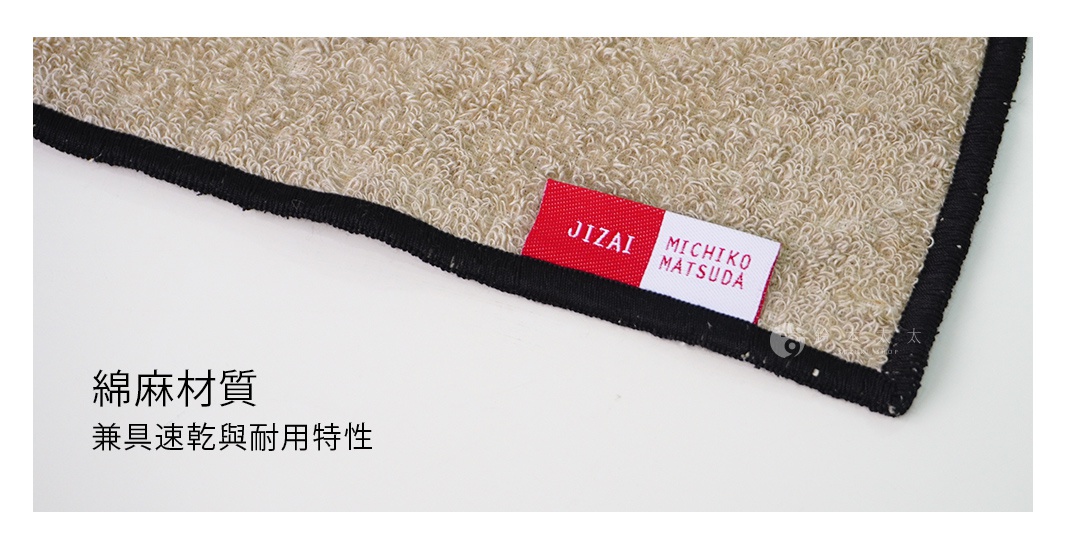 一張特寫圖片，展示了淺米色毛巾的紋理，邊緣有黑色滾邊。一條紅底白字的標籤，上面印有「JIZAI MICHIKO MATSUDA」字樣。文字區域顯示「棉麻材質 兼具速乾與耐用特性」。