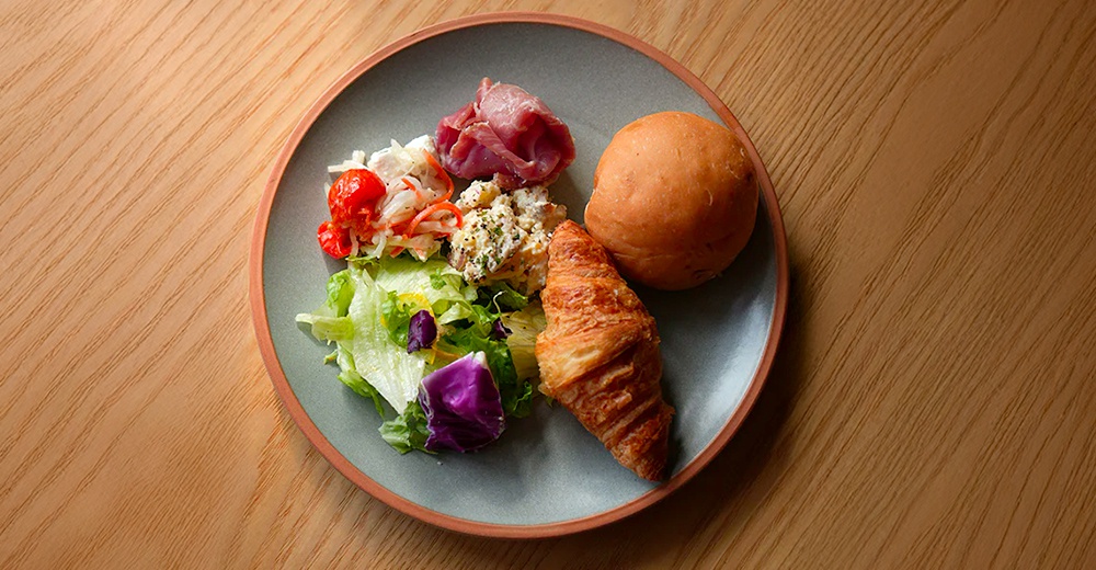 灰綠色陶瓷主菜盤，內有新鮮沙拉、火腿薄片、馬鈴薯沙拉、可頌麵包和圓形麵包，盤緣為橘紅色。