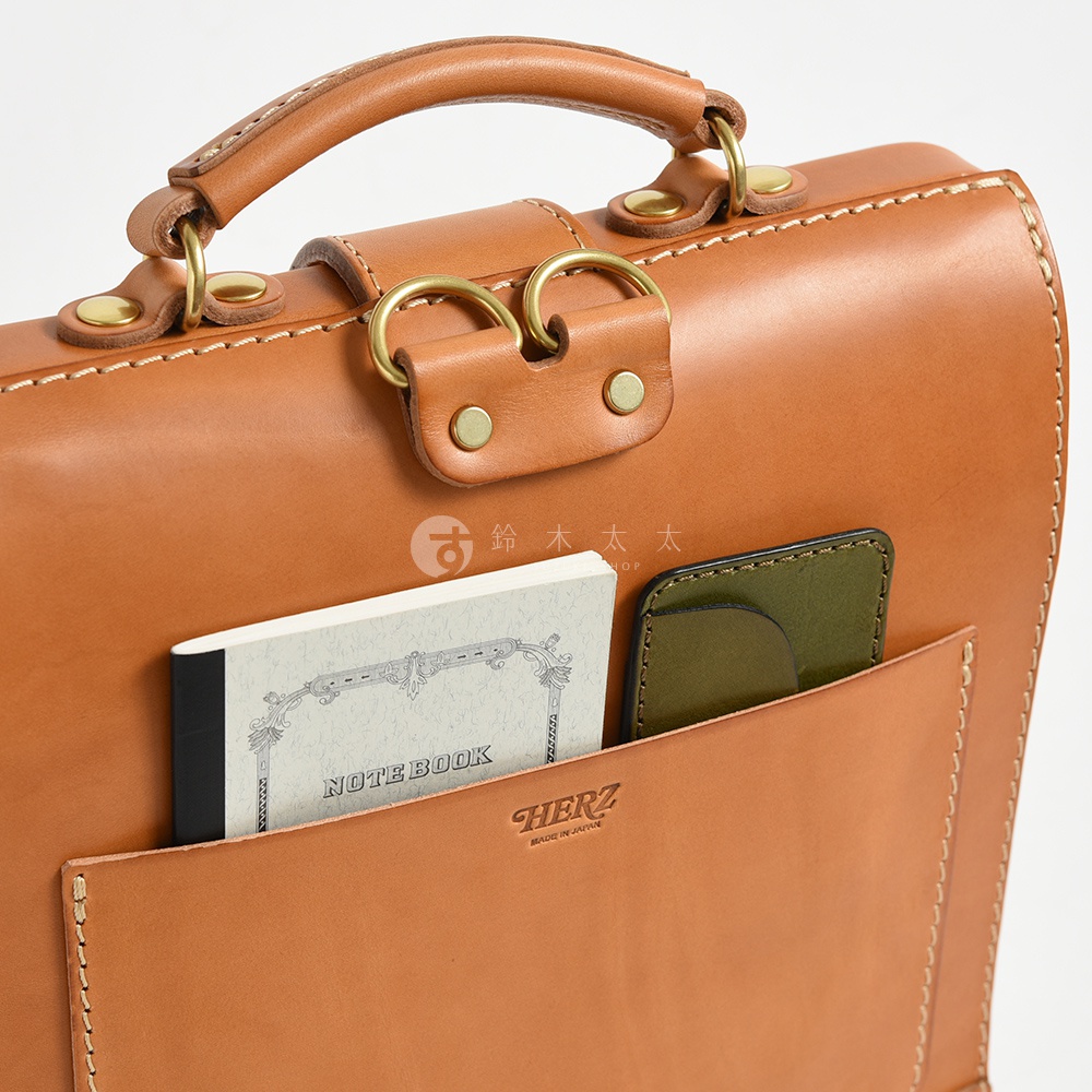 職人手作皮革杜勒斯商務後背包，棕褐色皮革材質，設有提把、金屬扣環及外部口袋，展示一本印有「NOTE BOOK」字樣的筆記本與一個綠色卡夾。
