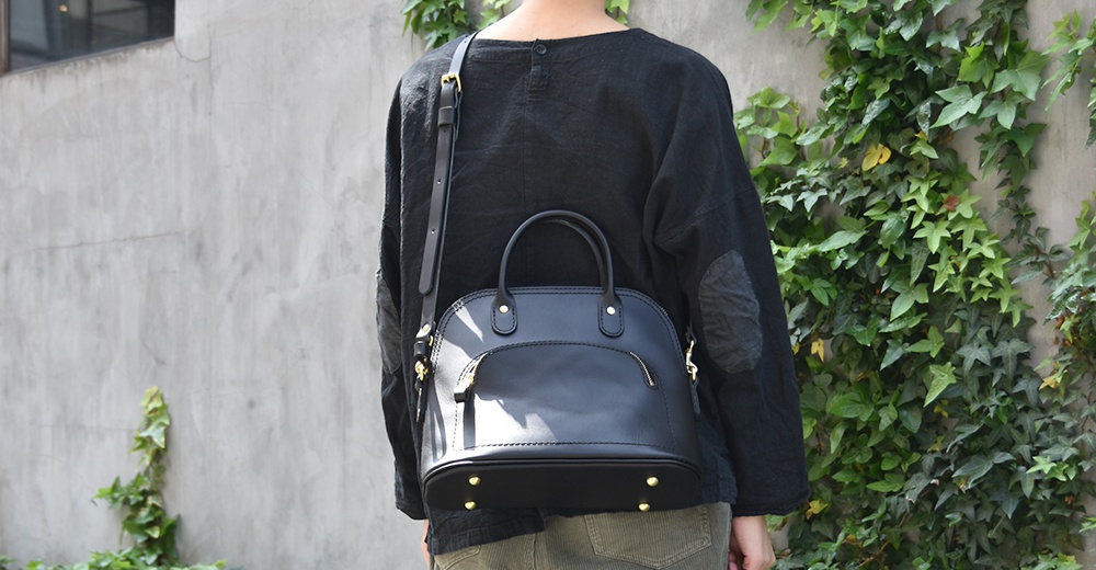 職人手作黑色皮革經典貝殼手提包，設有可拆卸肩帶、頂部提把和拉鍊前袋，底部有金屬腳釘。