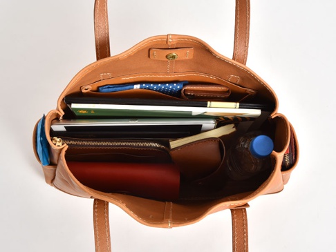 職人手作皮革橫式肩背托特包，棕色皮革材質，包內裝有平板電腦、筆記本、錢包、水瓶和藍色印花布料。