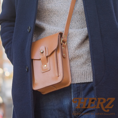 棕褐色皮革製作的方形斜背包，配有可調節的肩帶和金屬扣環。包包正面有縫線裝飾和一個翻蓋設計，透過按扣固定。