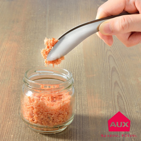 一只手持不鏽鋼夾，夾取罐中的鮭魚鬆，罐子放在木紋桌面上。