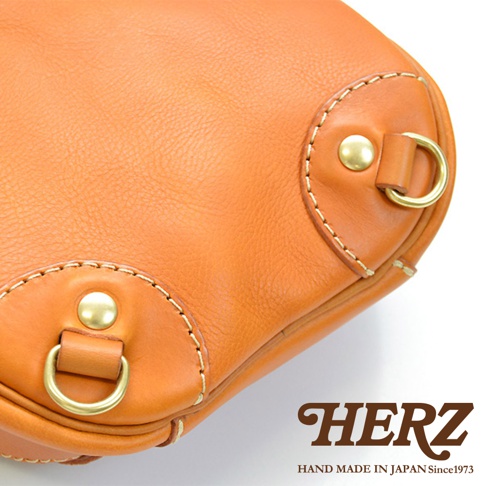 職人手作皮革斜背包，橘棕色皮革，有金色D形環和鉚釘裝飾，搭配米色縫線，展現精緻工藝。