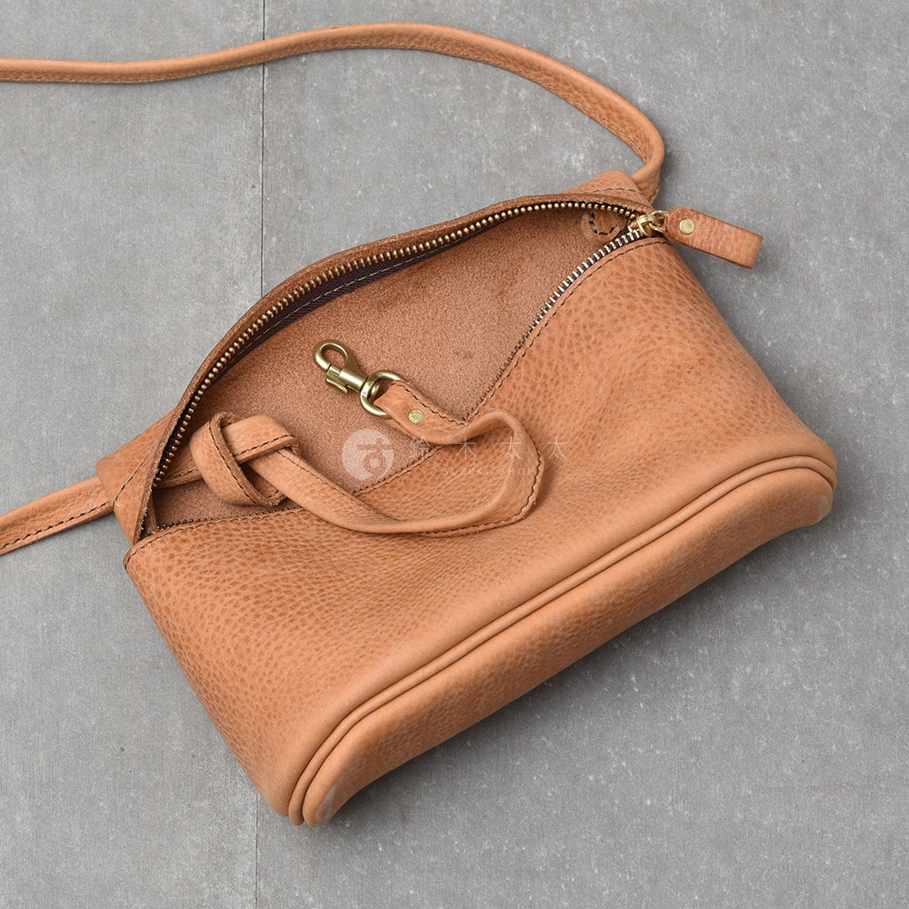 一個淺棕色皮革製成的斜背包，包身帶有紋理，開口處為拉鍊設計，並有一個金屬鉤扣。